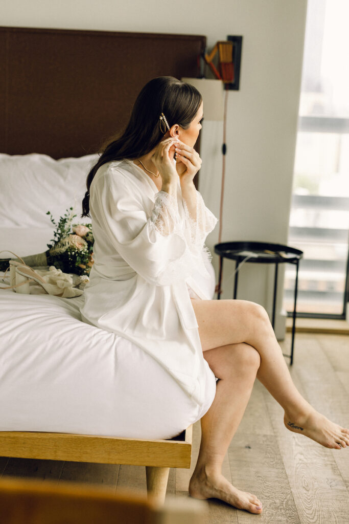 A woman, sitting on a white, hotel bed, puts in an earring to get ready for her wedding ceremony.