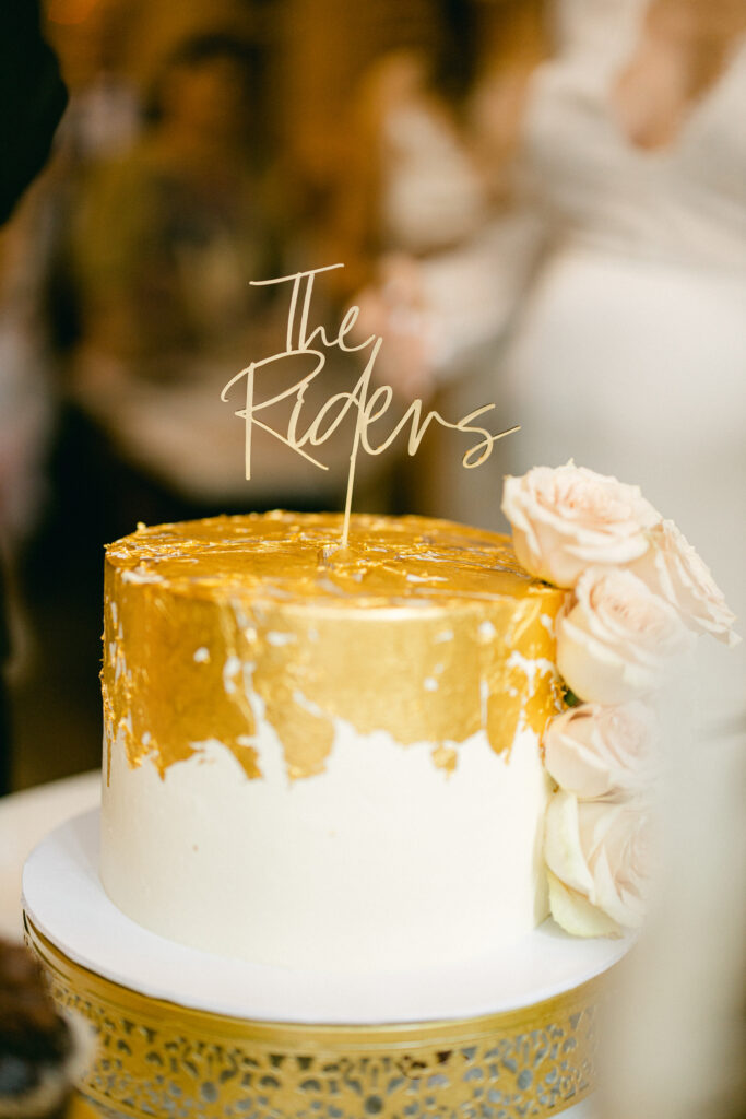 A white wedding cake with gold foil decor, pink roses trailing the side, and a gold wedding topper that reads "The Riders."