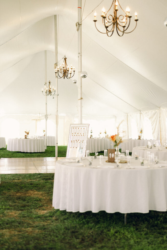 The wedding reception decor with white linens, colorful florals, and brass chandeliers under a covered tent.
