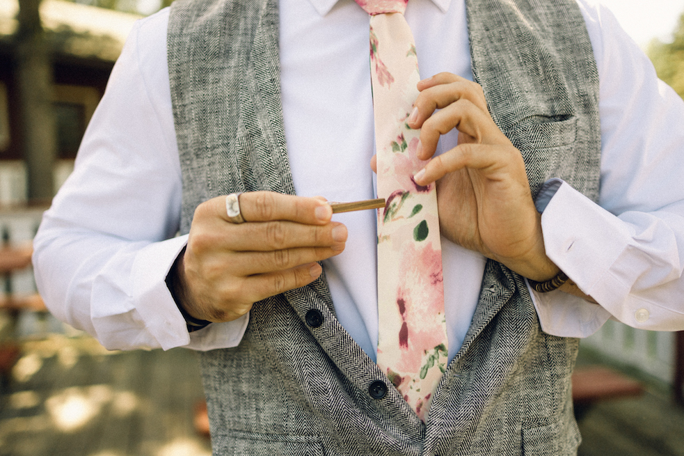 The groom wearing a pink, floral tie and putting on a wooden tie clip.