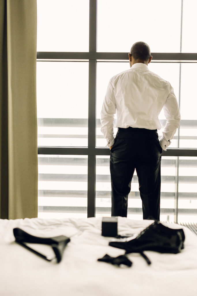 The groom looks out into the New York City skyline from the hotel windows with his shoes, cologne, and bow tie in focus. 