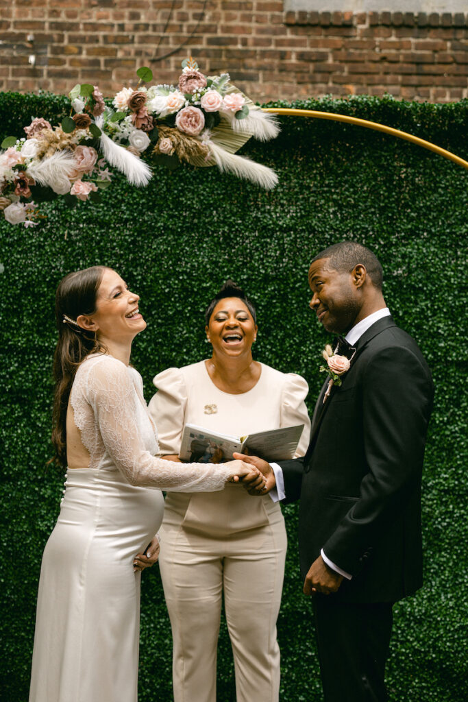 The wedding officiant makes a joke and laughs with the bride and groom at the altar, standing in front of greenery beneath a gold and floral arch at Levante NYC's outdoor courtyard space.