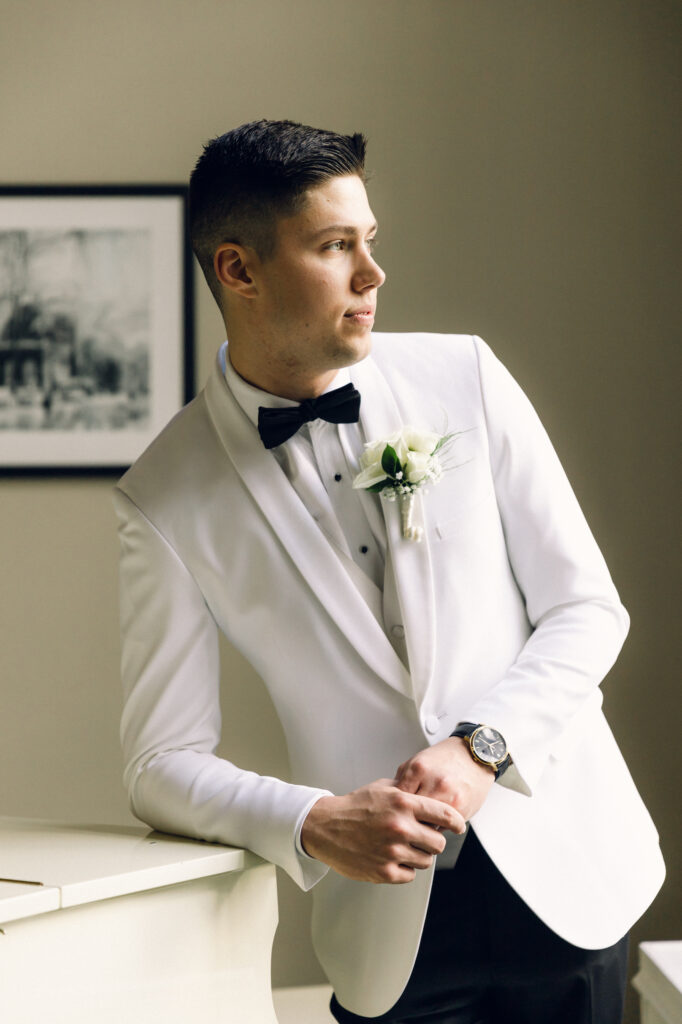 The groom wearing a white tuxedo jacket, black tuxedo pants, and a black bow tie.