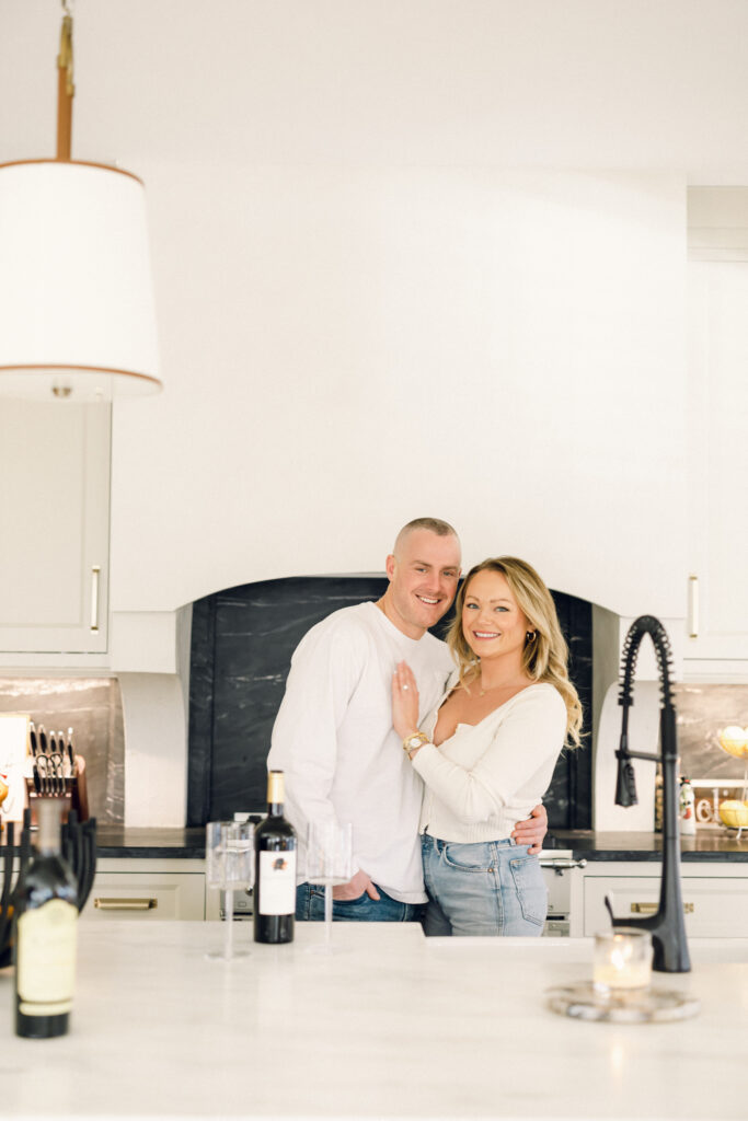 at-home engagement session with the couple dressed in white, smiling in the kitchen while drinking wine.