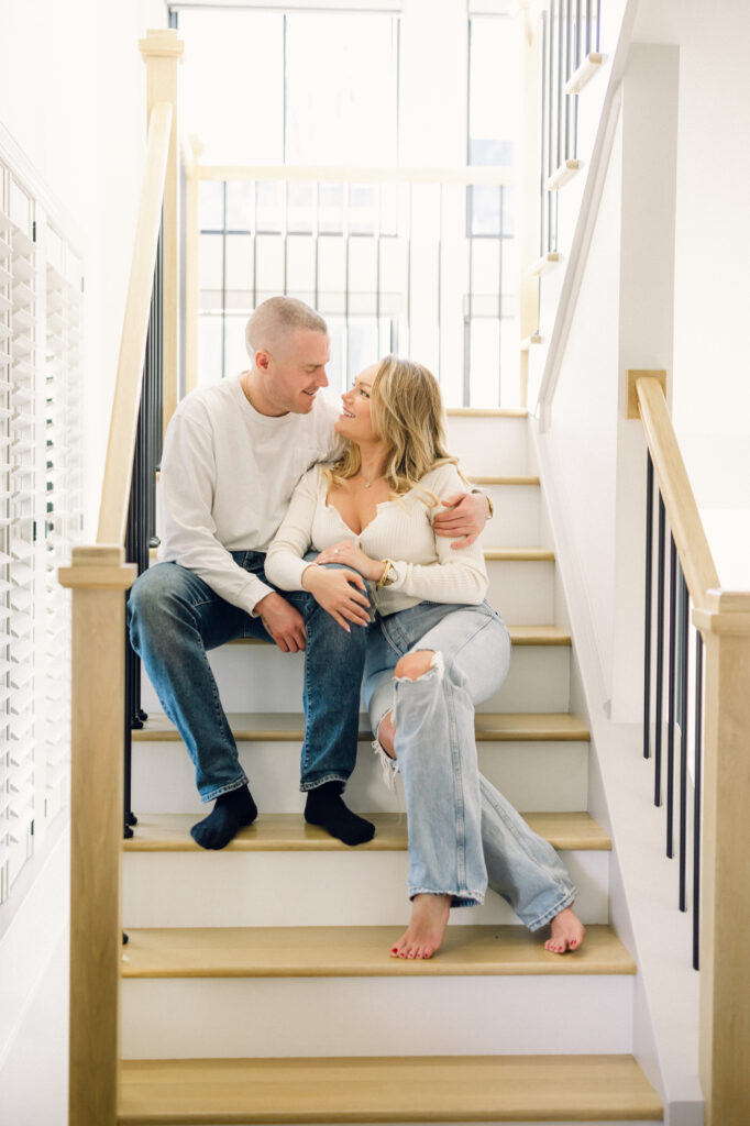 at-home engagement session with the couple dressed in white, sitting on their staircase, looking at one another smiling. 