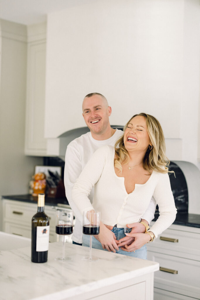 Upstate New York at-home engagement session with the couple dressed in white, smiling in the kitchen while drinking wine.
