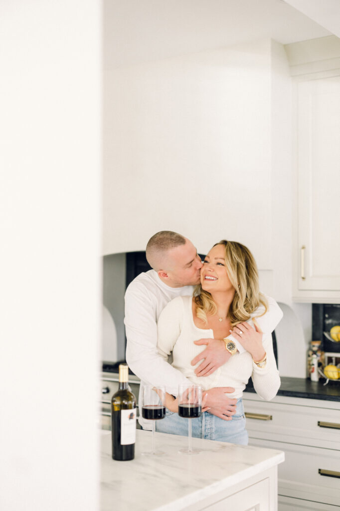 new York at-home engagement session with the couple dressed in white, smiling in the kitchen while drinking wine.