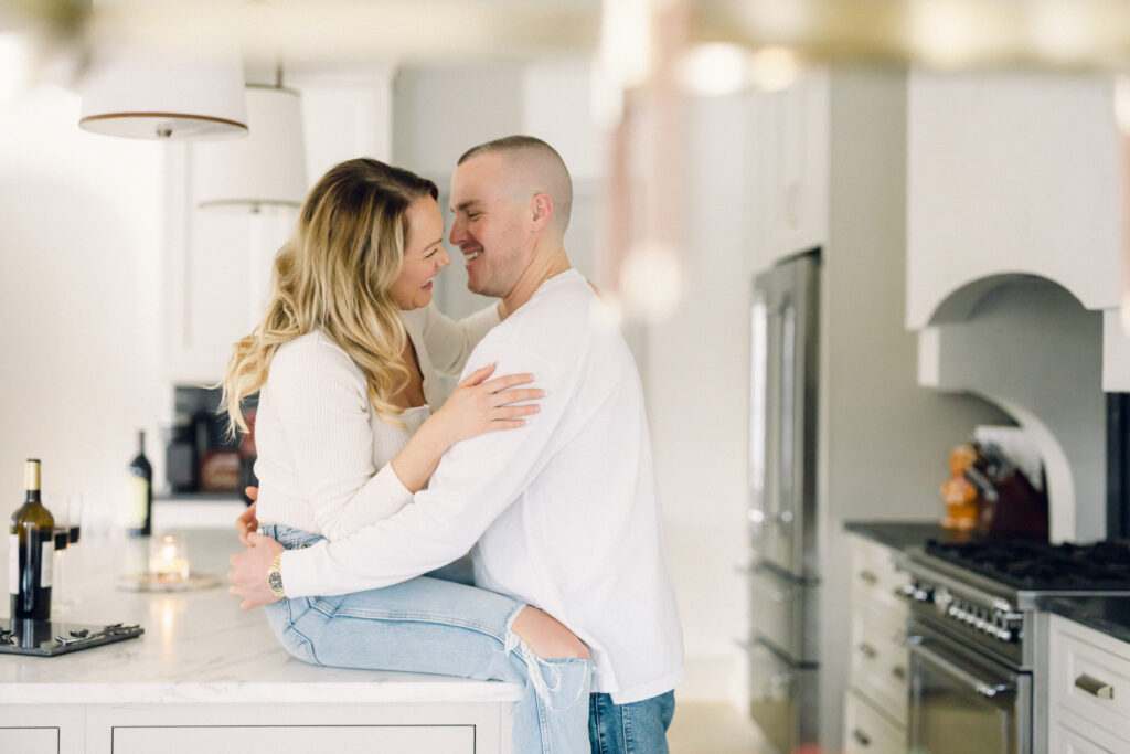 New York City at-home engagement session with the couple dressed in white, smiling in the kitchen while drinking wine.