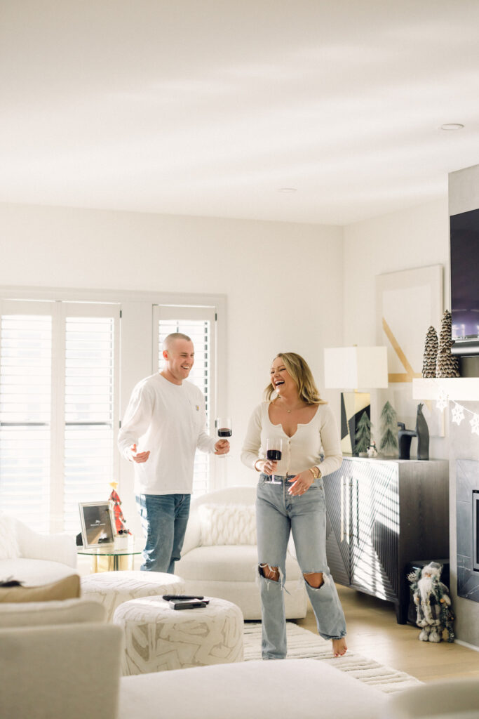 at-home engagement session with the couple dressed in white, dancing with wine in the living room for documentary-style photos.