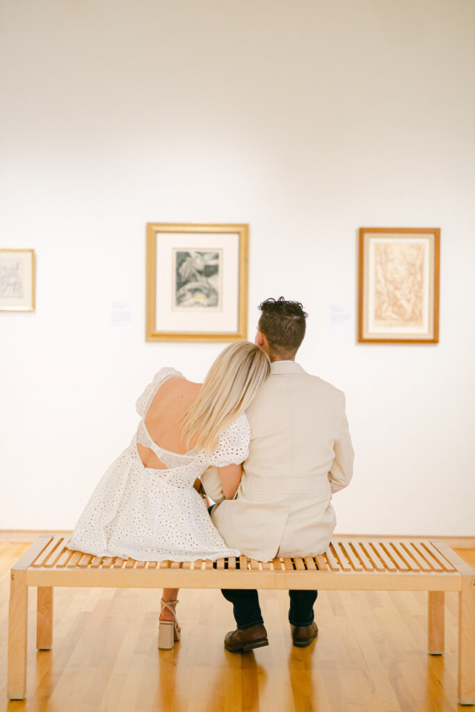 Couple looks at framed art in a museum while sitting on a bench.