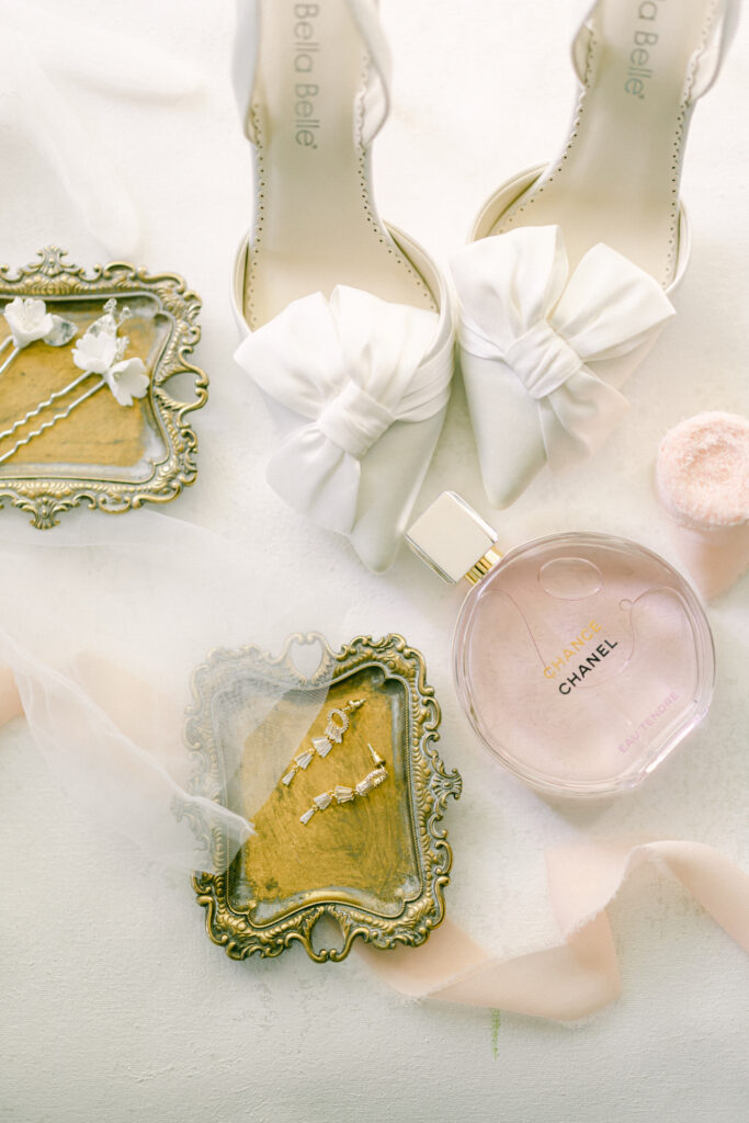 Detail wedding photography shot with a pink bottle of chanel perfume, white heels with satin bows, jewelry in a brass dish, white bow hairpins, and a pink ribbon draped through the view. 
