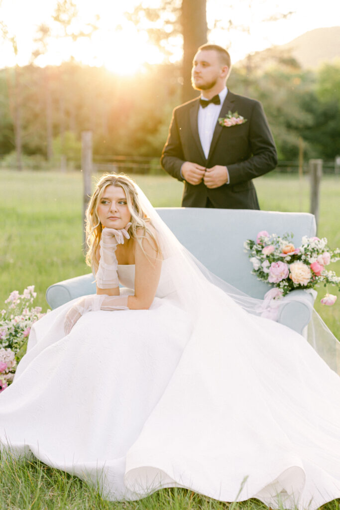 Bride looking into a meadow while sitting in a blue loveseat while pink florals and the groom behind her.