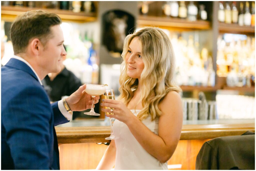 A bride and groom toast their drinks while standing at the bar