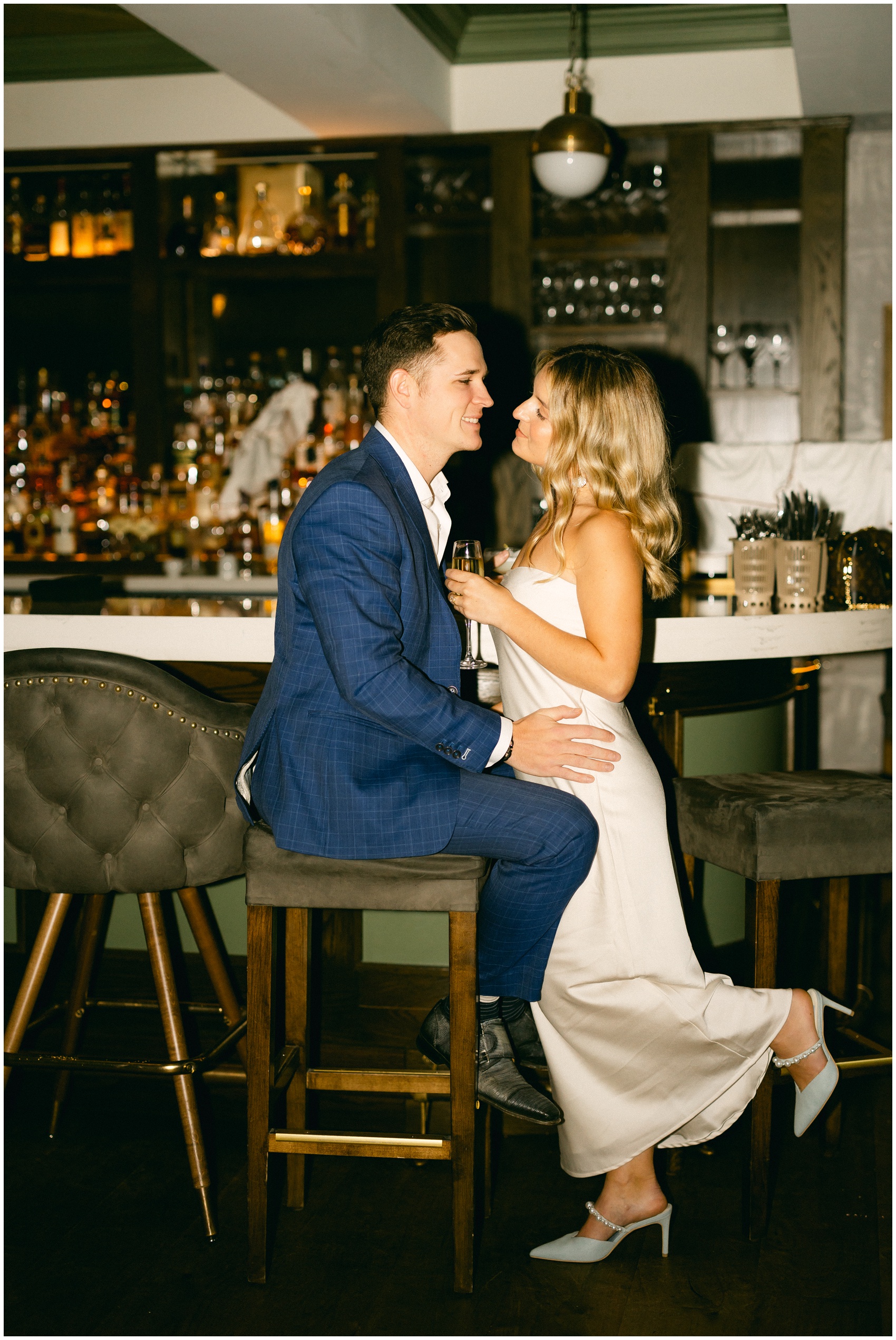 A bride leans into her groom while he sits on a bar stool at the bar with champagne