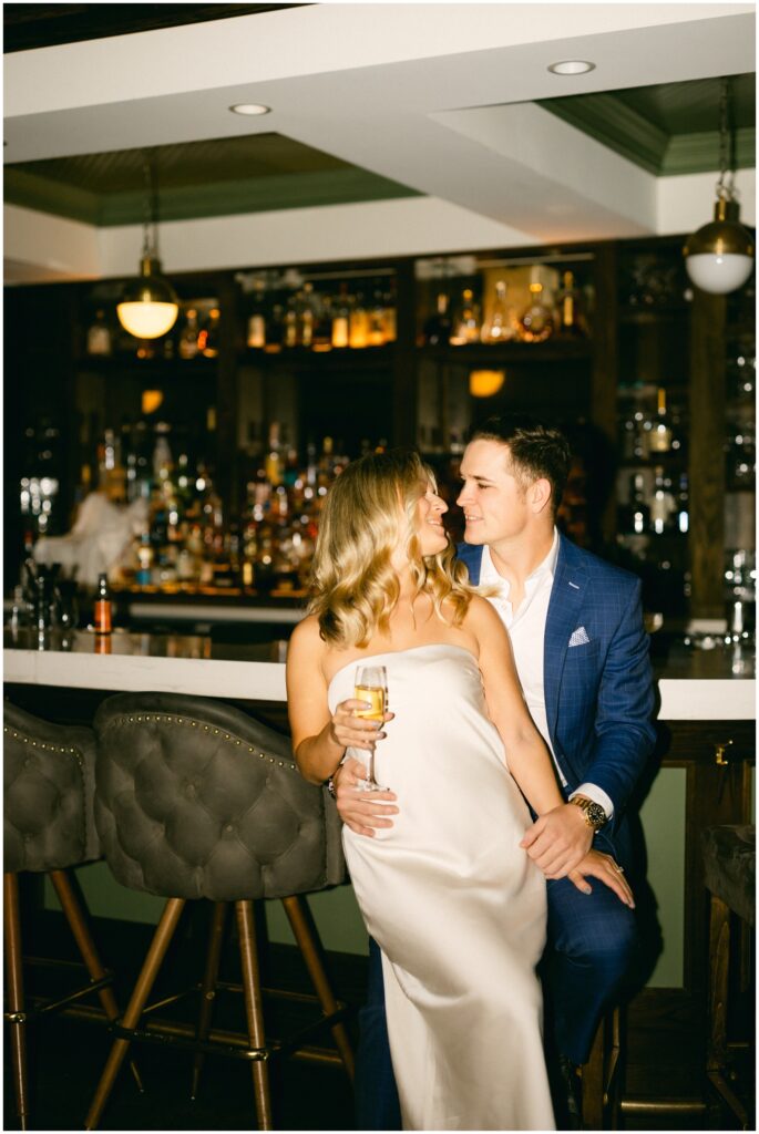 A bride sits in the lap of her groom at the bar as they lean in for a kiss