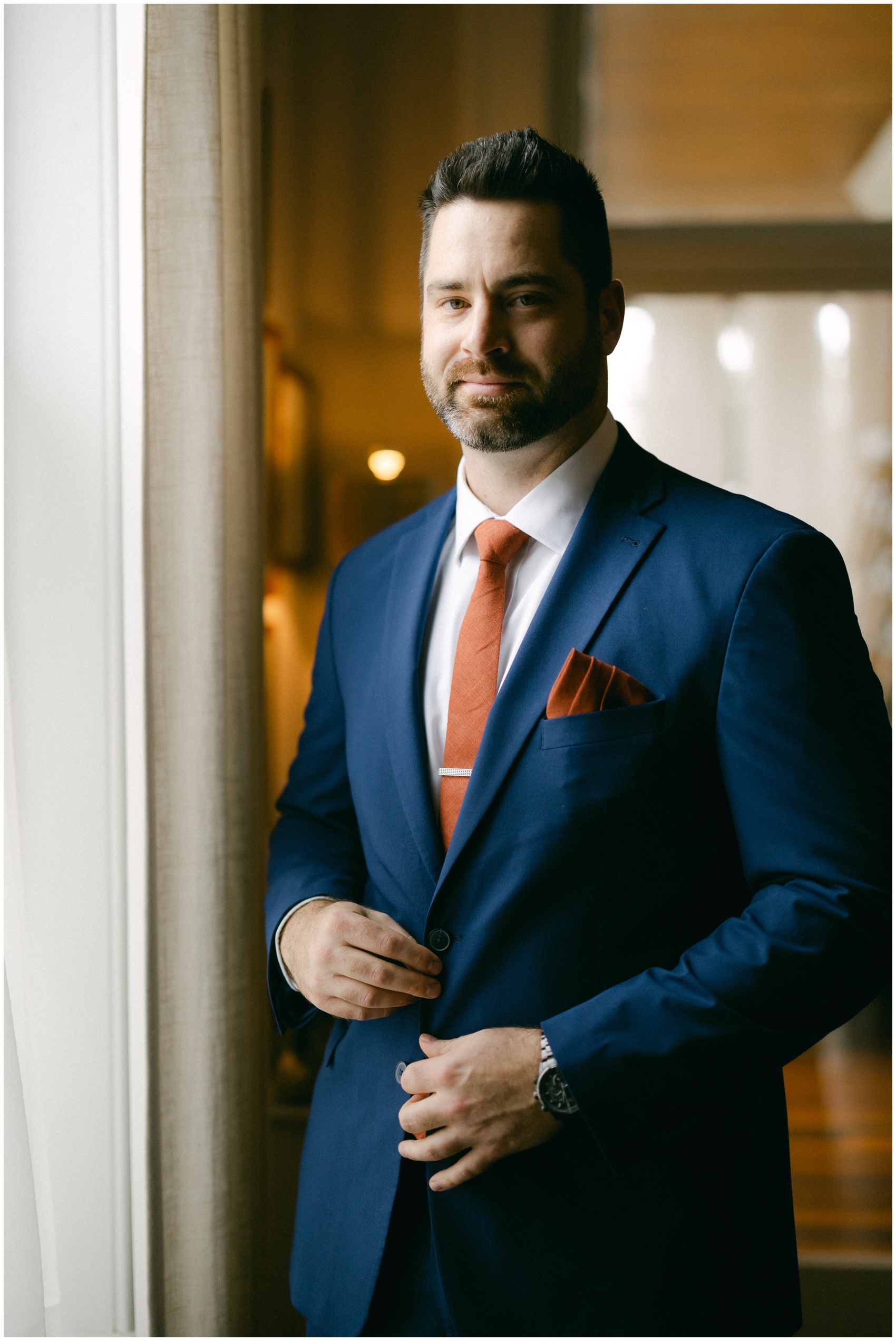 A groom in a blue suit stands in a window smiling with an orange tie