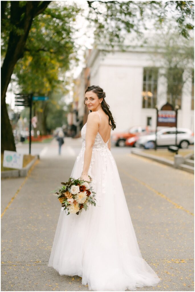 A bride smiles while walking on the sidewalk with her bouquet in her hand by her side