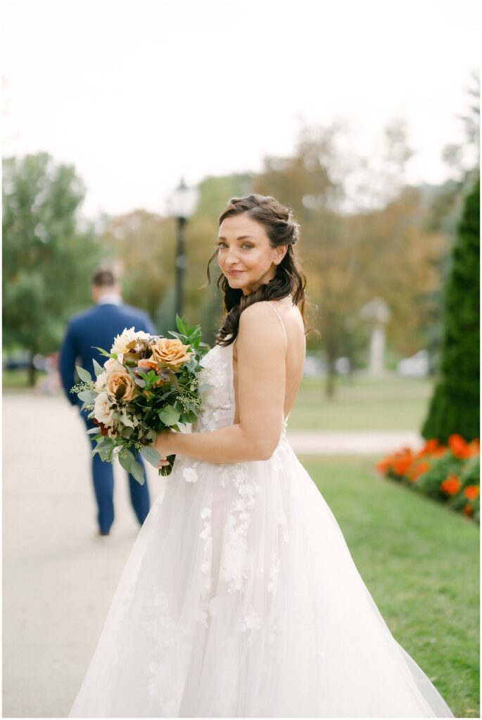 A bride smiles over her shoulder while holding her bouquet with her groom standing down the sidewalk