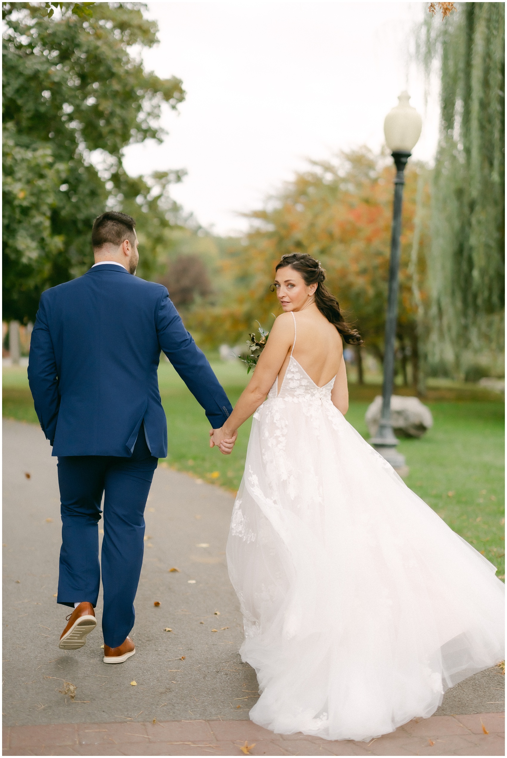 A bride looks over her shoulder while walking with her groom in a park sidewalk at the mansion of saratoga