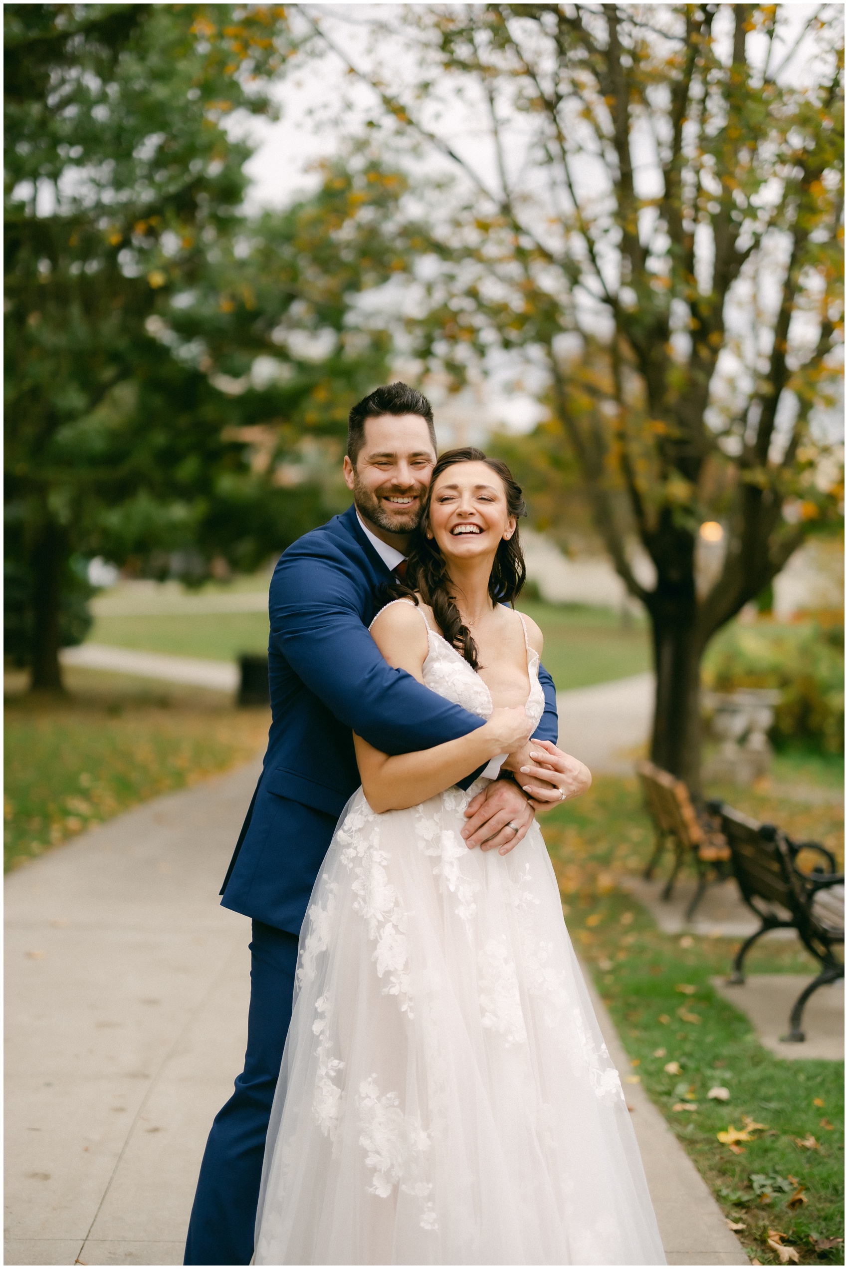 A happy newlywed couple hugs while walking in a park sidewalk and laughing together