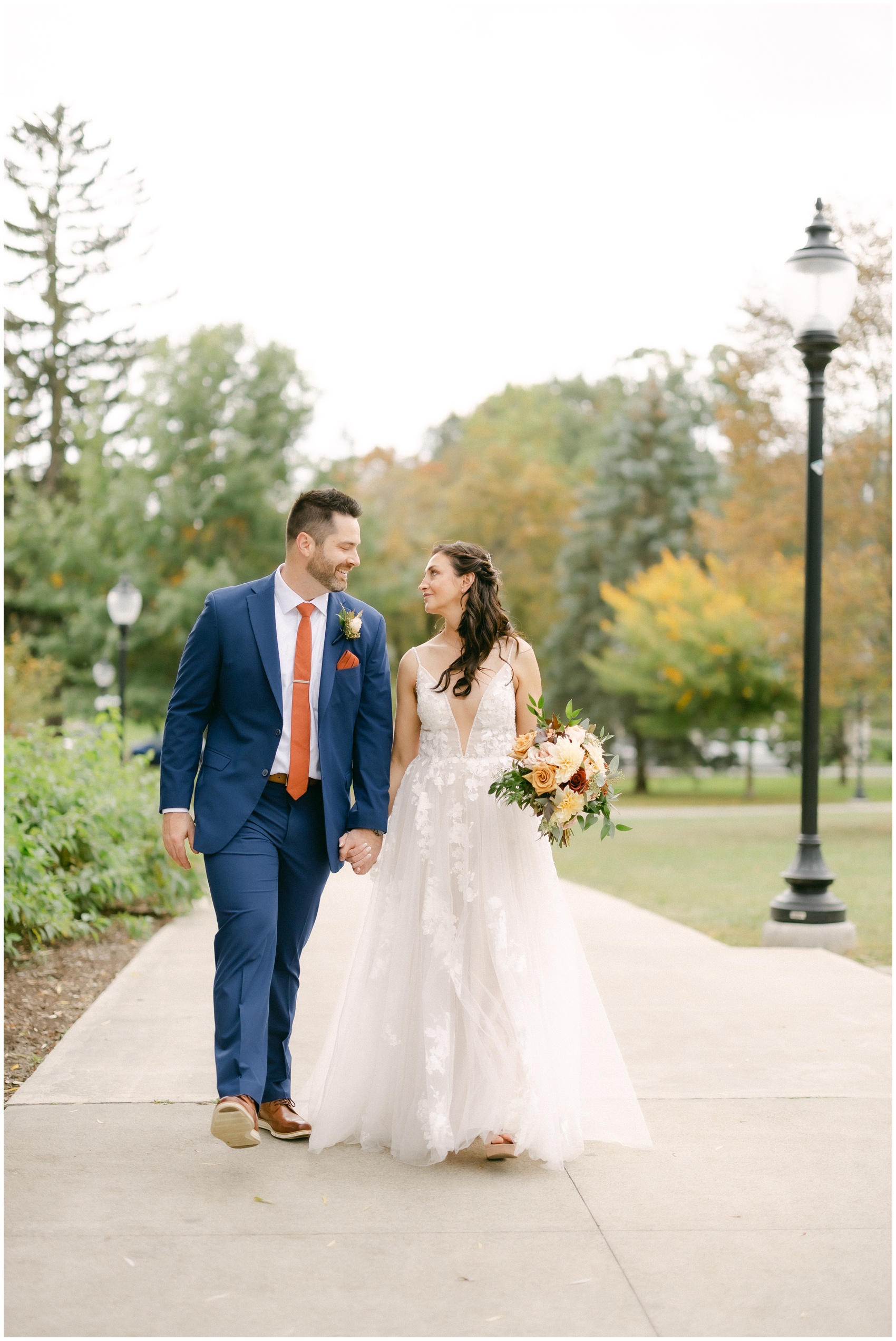 Newlyweds hold hands while walking on a park sidewalk and smiling at each other at the mansion of saratoga