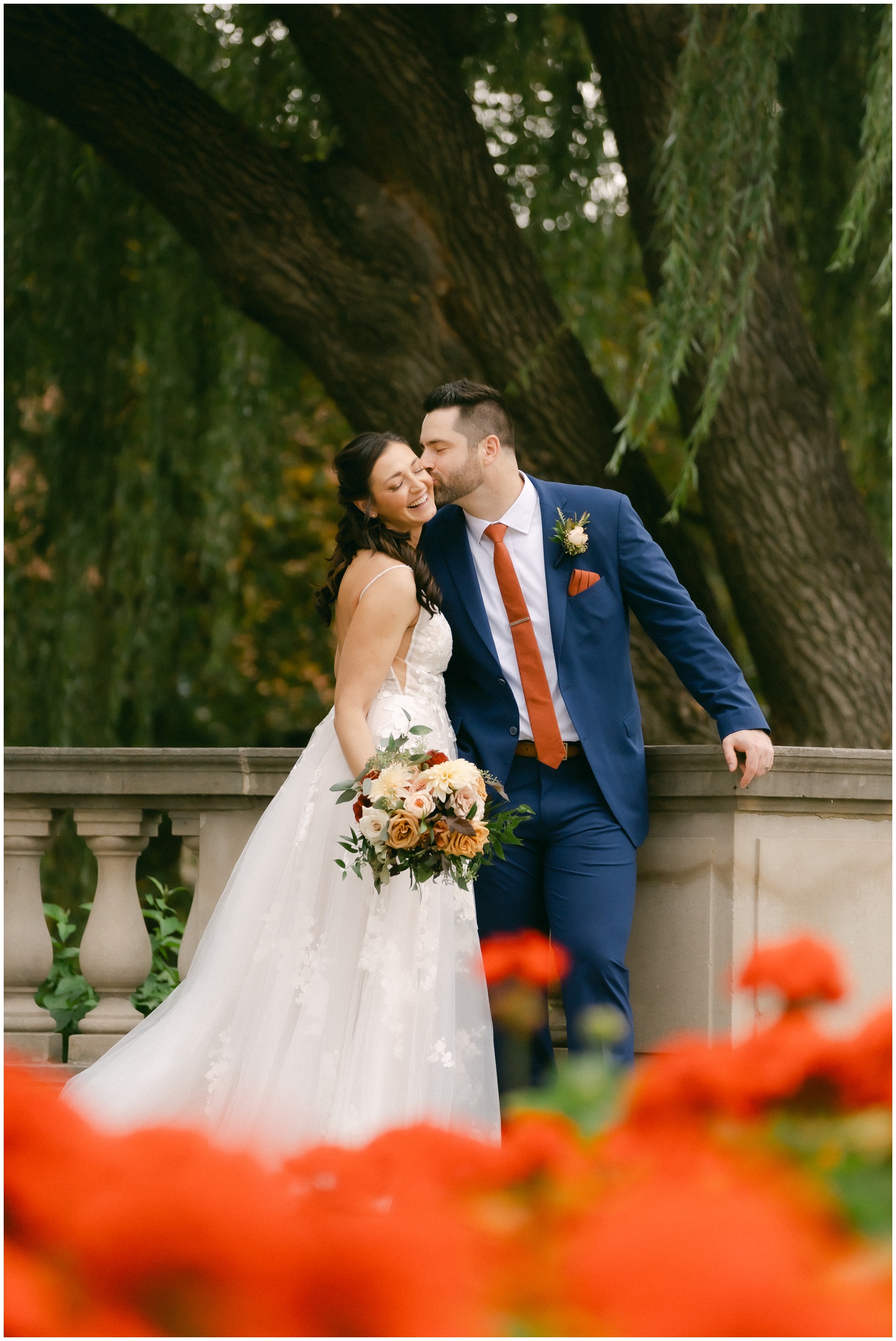 A bride smiles while being kissed by her groom in a blue suit while leaning on a garden railing under a willow tree at the mansion of saratoga