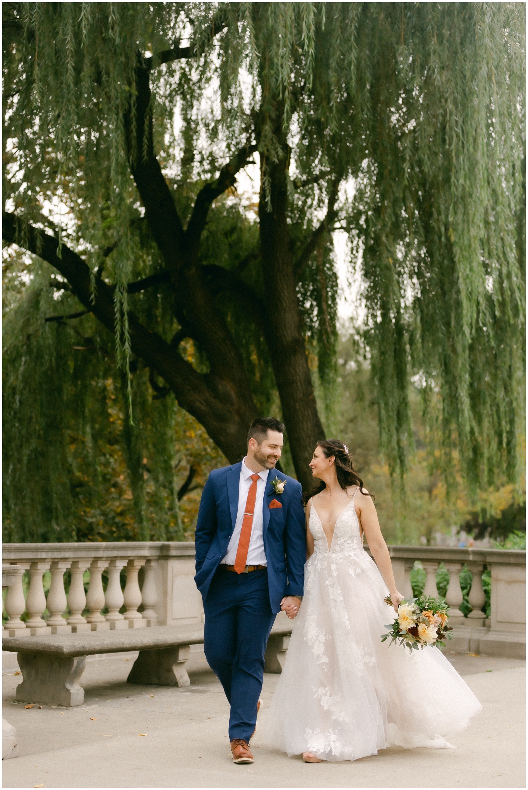 Newlyweds hold hands walking under a willow tree in the gardens at the mansion of saratoga