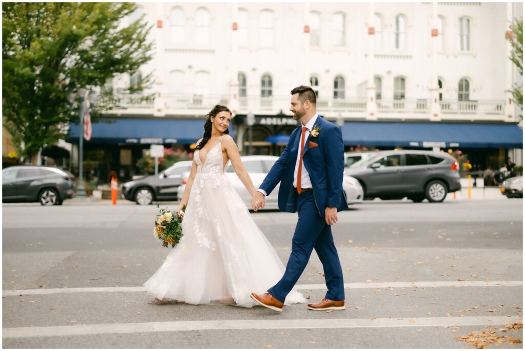 A bride looks back to her groom as they hold hands walking across a street at the mansion of saratoga