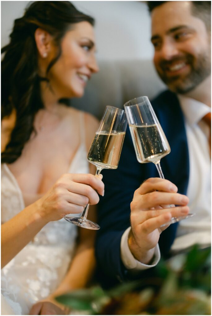 Newlyweds cheers their champagne glasses while sitting on a couch and smiling at each other